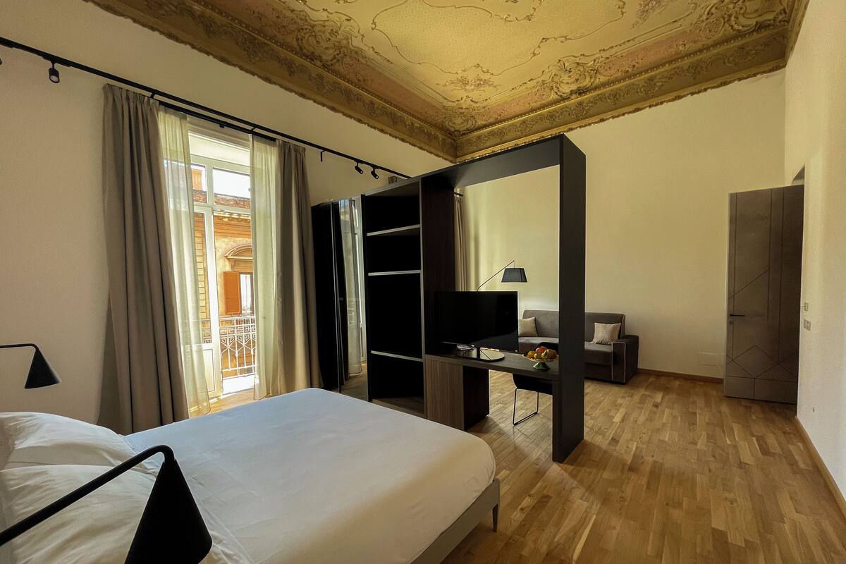 Una camera d'albergo con un letto, una scrivania con televisore, un armadio e vista sul balcone esterno.