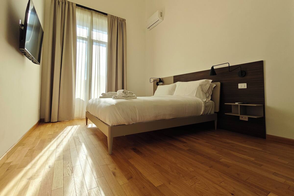 Una moderna camera da letto con un letto, una TV a schermo piatto montata a parete, una finestra con tende e una mensola a incasso.