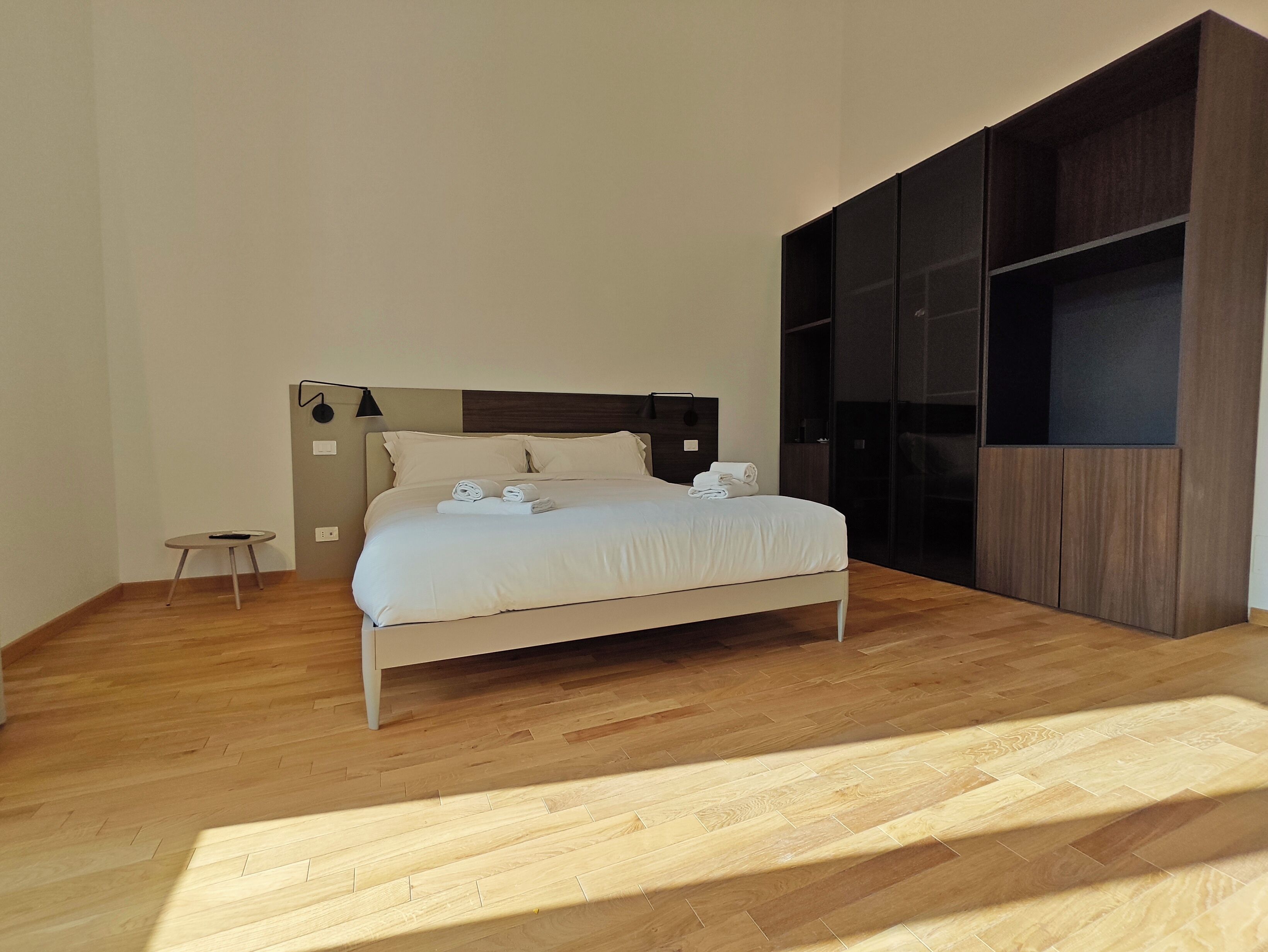A modern bedroom with a wooden floor, a bed with white linens, a small bedside table, and a built-in wardrobe.