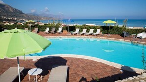 En swimmingpool med solstolar och parasoller, omgiven av en innergård med tegelbeläggning och utsikt över havet och bergen.