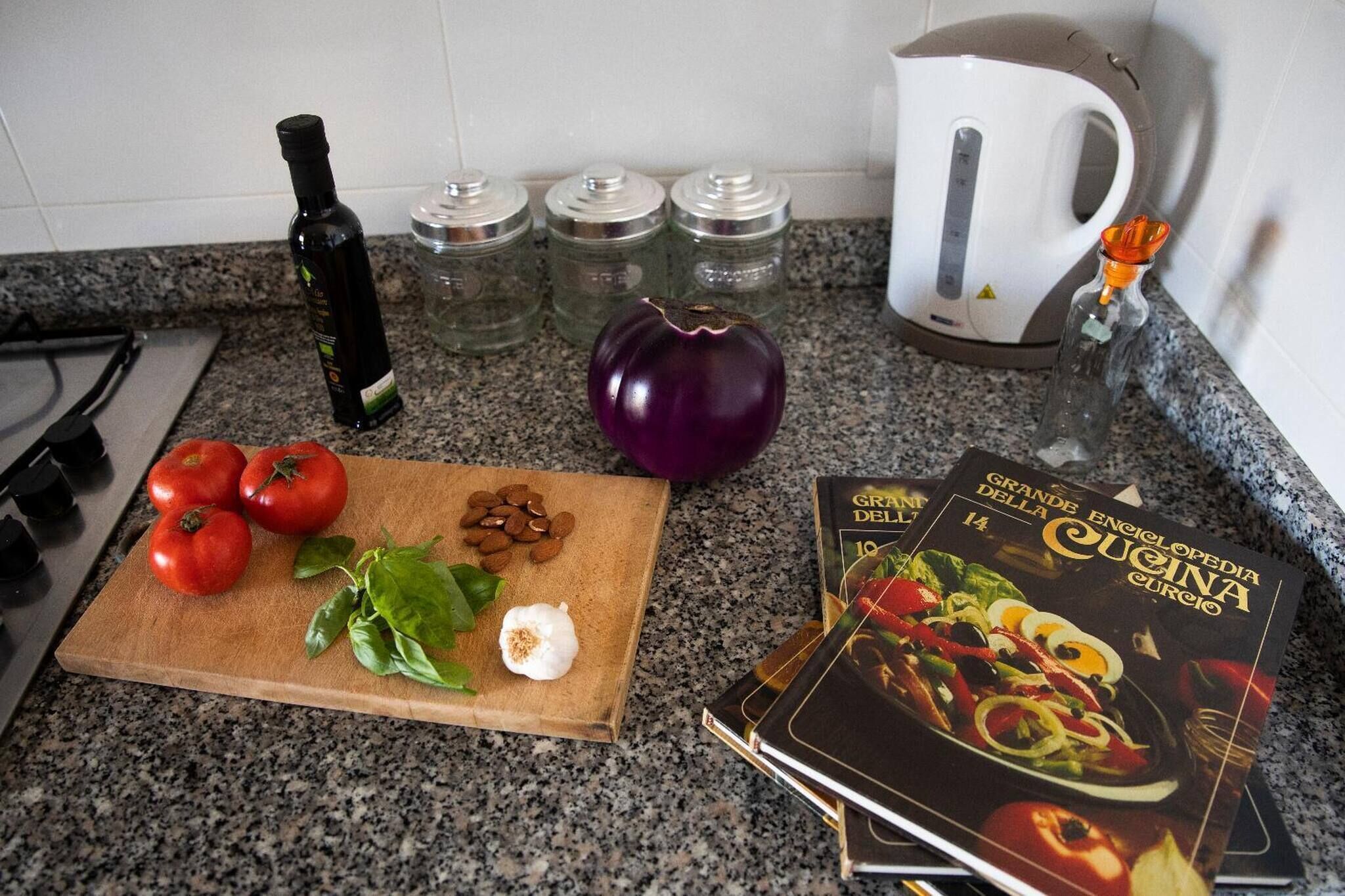 A kitchen counter with tomatoes, a purple eggplant, a bottle of olive oil, salt and pepper shakers, a kettle, and a cookbook.