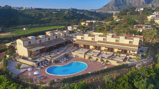 Ett semesterhotell med en swimmingpool omgiven av solstolar och parasoller, med kullar och grönskande vegetation som bakgrund.