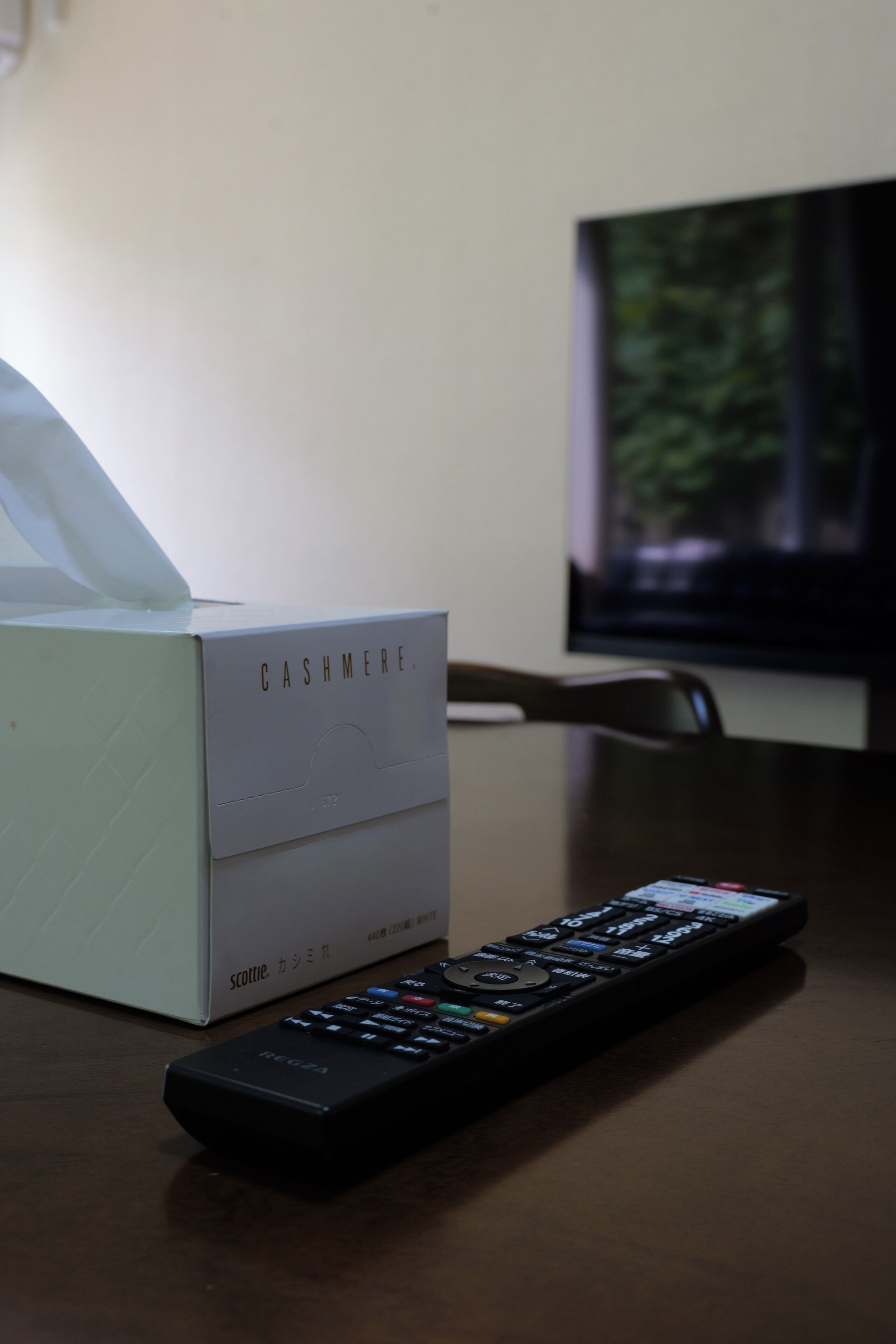 Un control remoto, una caja de pañuelos y un televisor sobre una mesa de madera.
