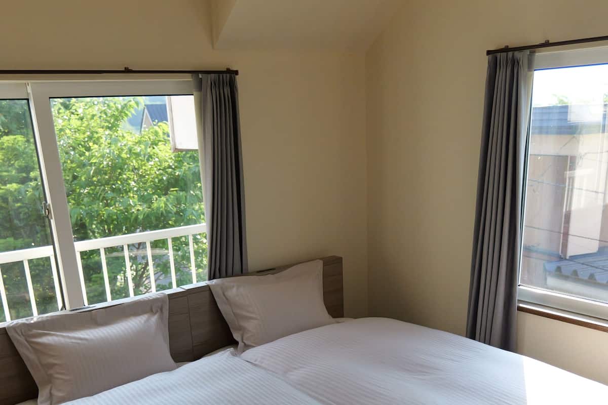 Una cama grande con ropa blanca, dos ventanas con cortinas y vista a la vegetación exterior.
