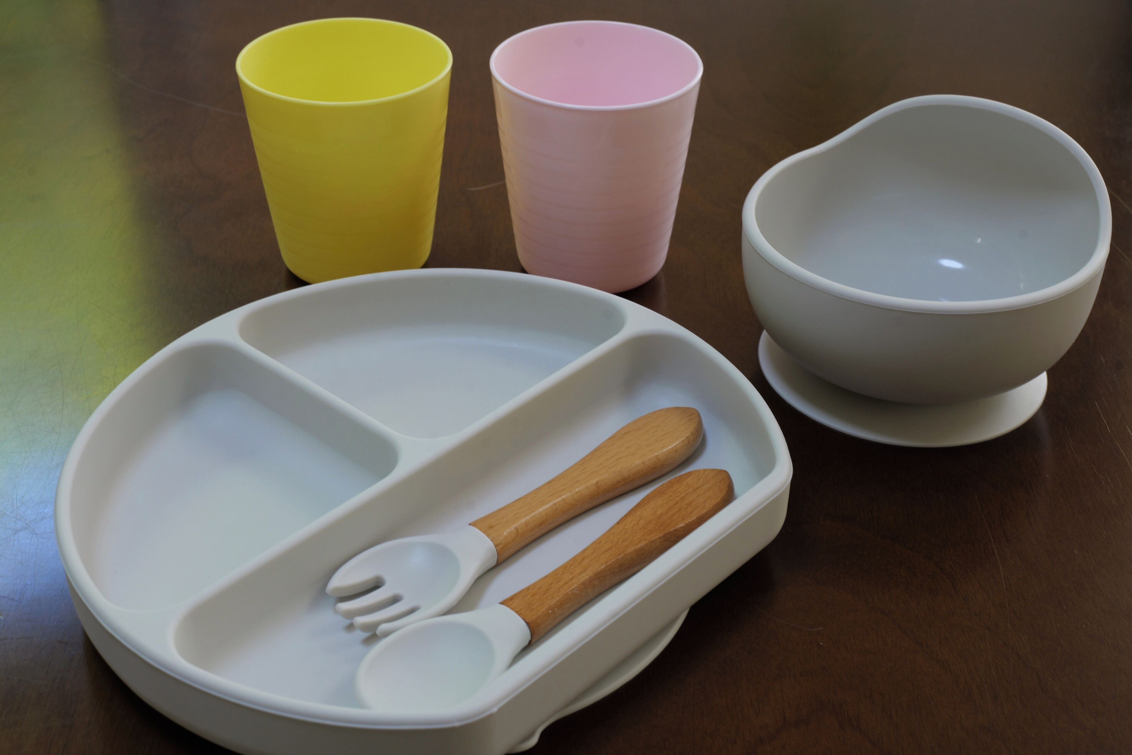A white tray with a fork and spoon, a yellow cup, a pink cup, and a white bowl on a wooden table.