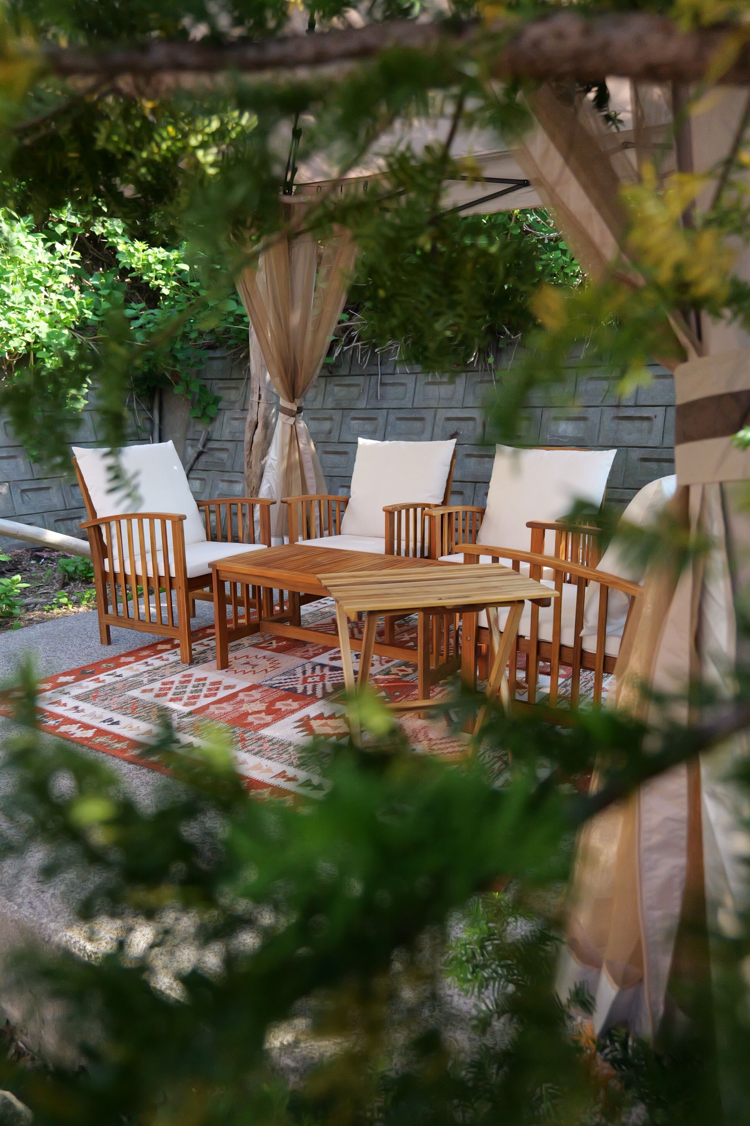 A wooden dining table with chairs, a patterned rug, and a backdrop of a stone wall and greenery.