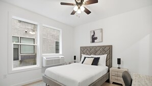 Un dormitorio con cama, mesita de noche, silla, ventilador de techo y ventana con persianas.