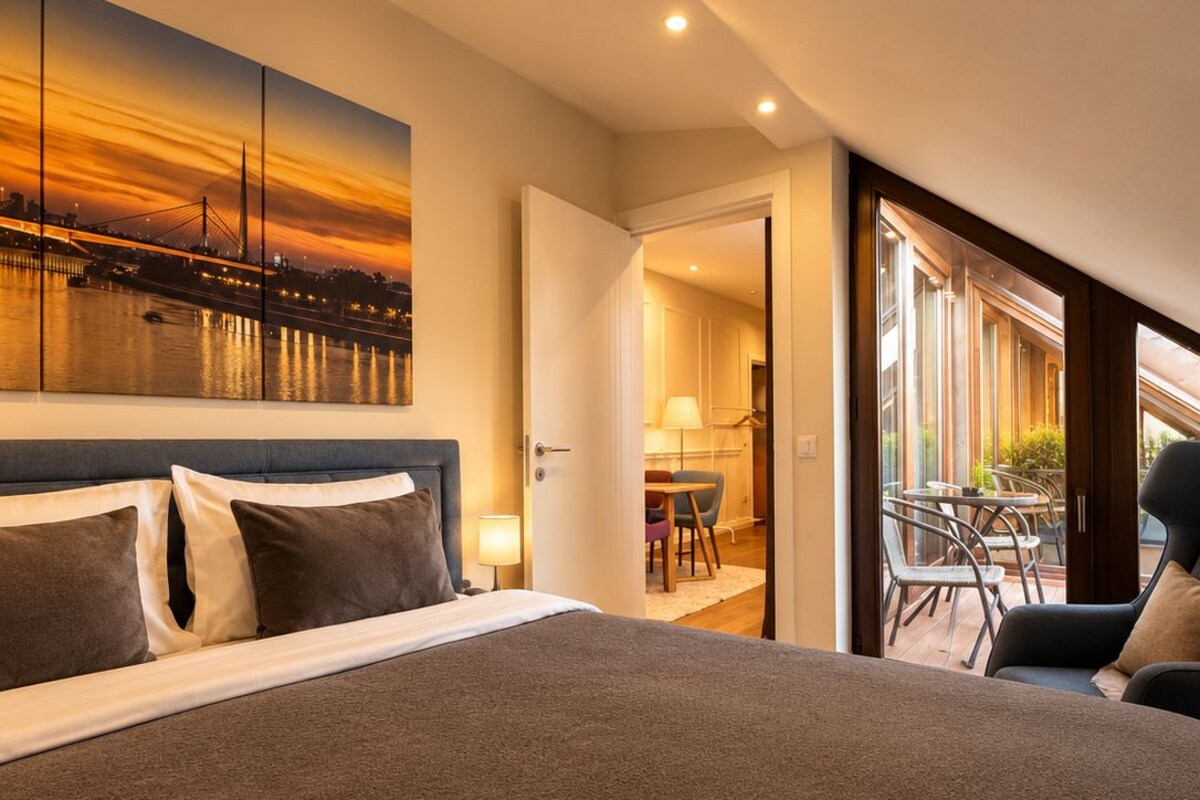 A hotel room with a large bed, a cityscape view, and a balcony with outdoor seating.