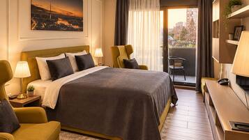 Habitación de hotel con cama, dos sillones, un escritorio, una silla, ventana con cortinas, un cuadro en la pared y balcón con vistas.