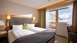 A hotel room with a large bed, wooden headboard, bedside lamps, a chair, a window with a mountain view, and a wooden desk with a book and phone. - Alpensporthotel Mutterberg (Neustift Im Stubaital)