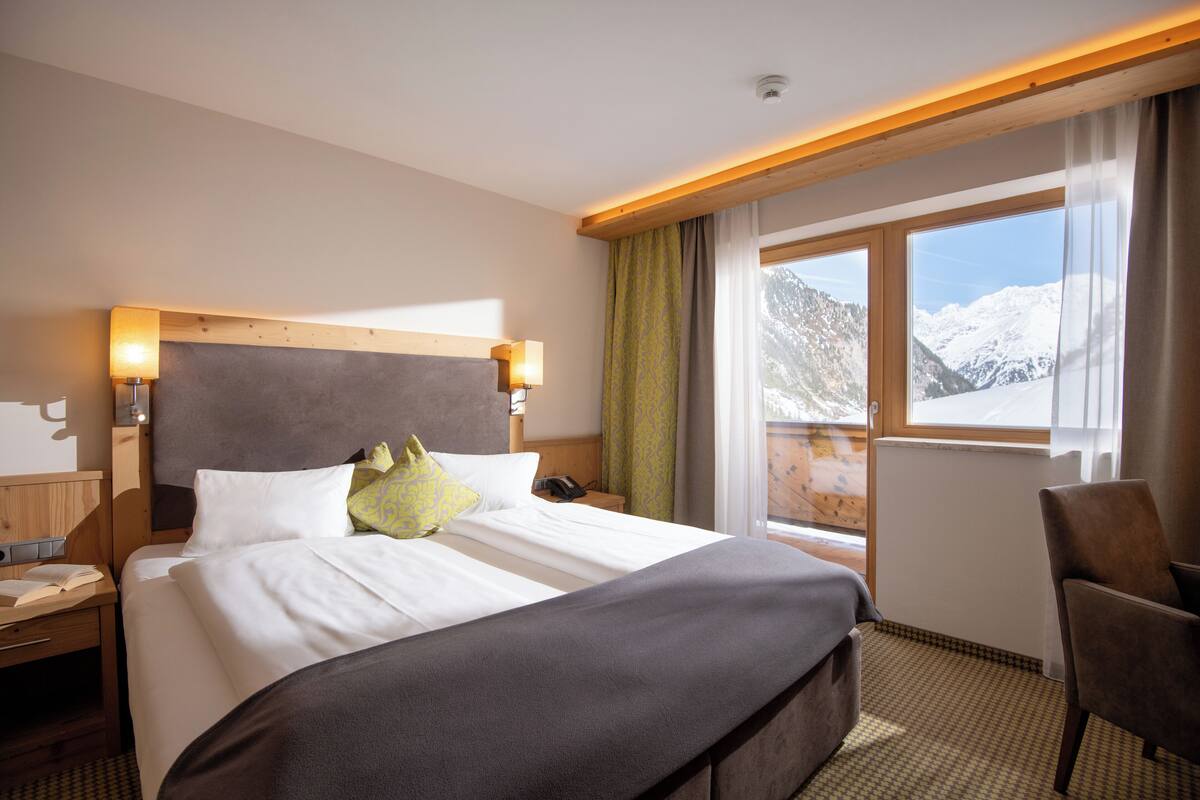 Une chambre d’hôtel avec un grand lit, une tête de lit en bois, des lampes de chevet, une chaise, une fenêtre donnant sur la montagne et un bureau en bois avec un livre et un téléphone.