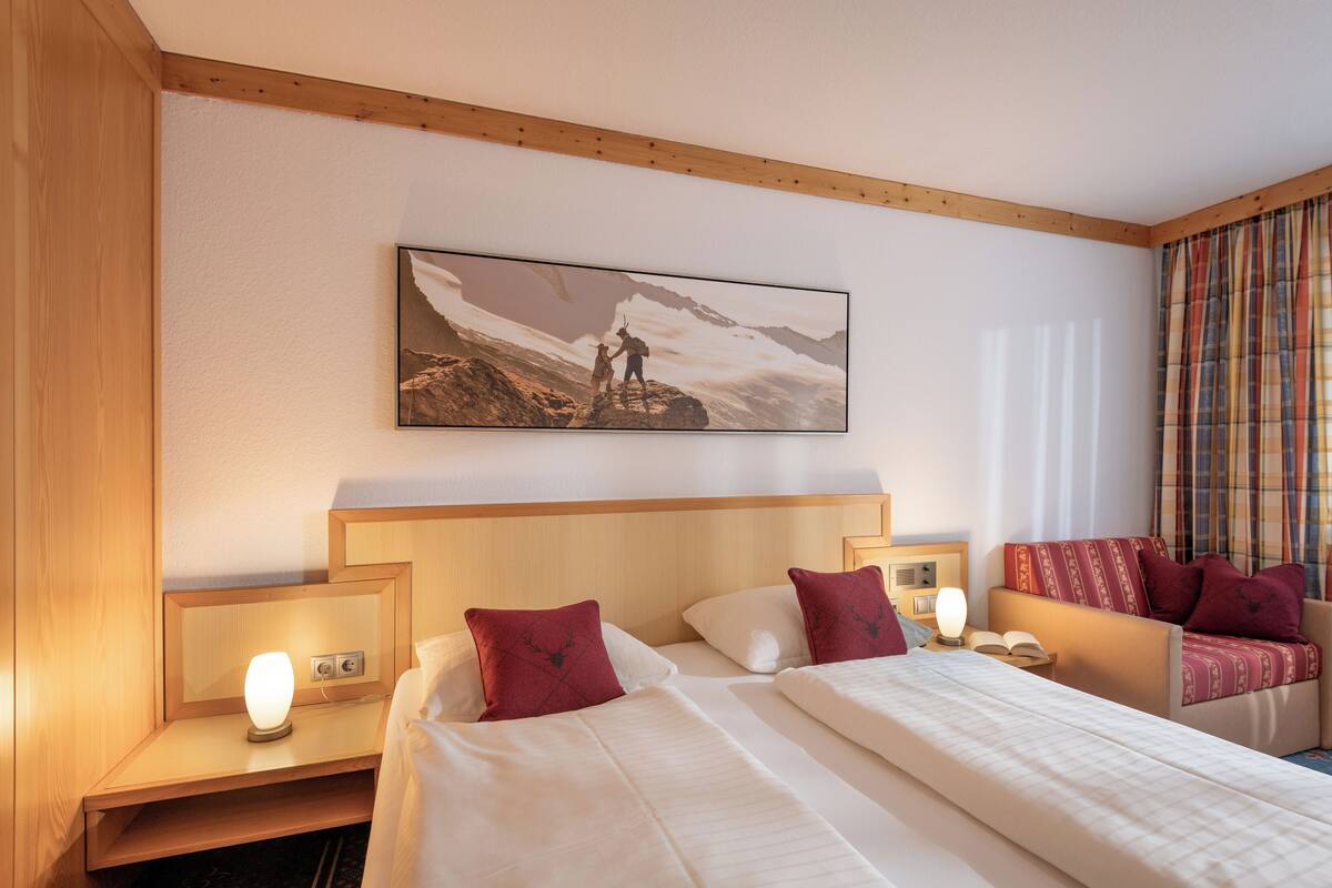 Une chambre d’hôtel avec un grand lit, deux tables de chevet, une commode, un canapé et un tableau représentant un paysage de montagne.