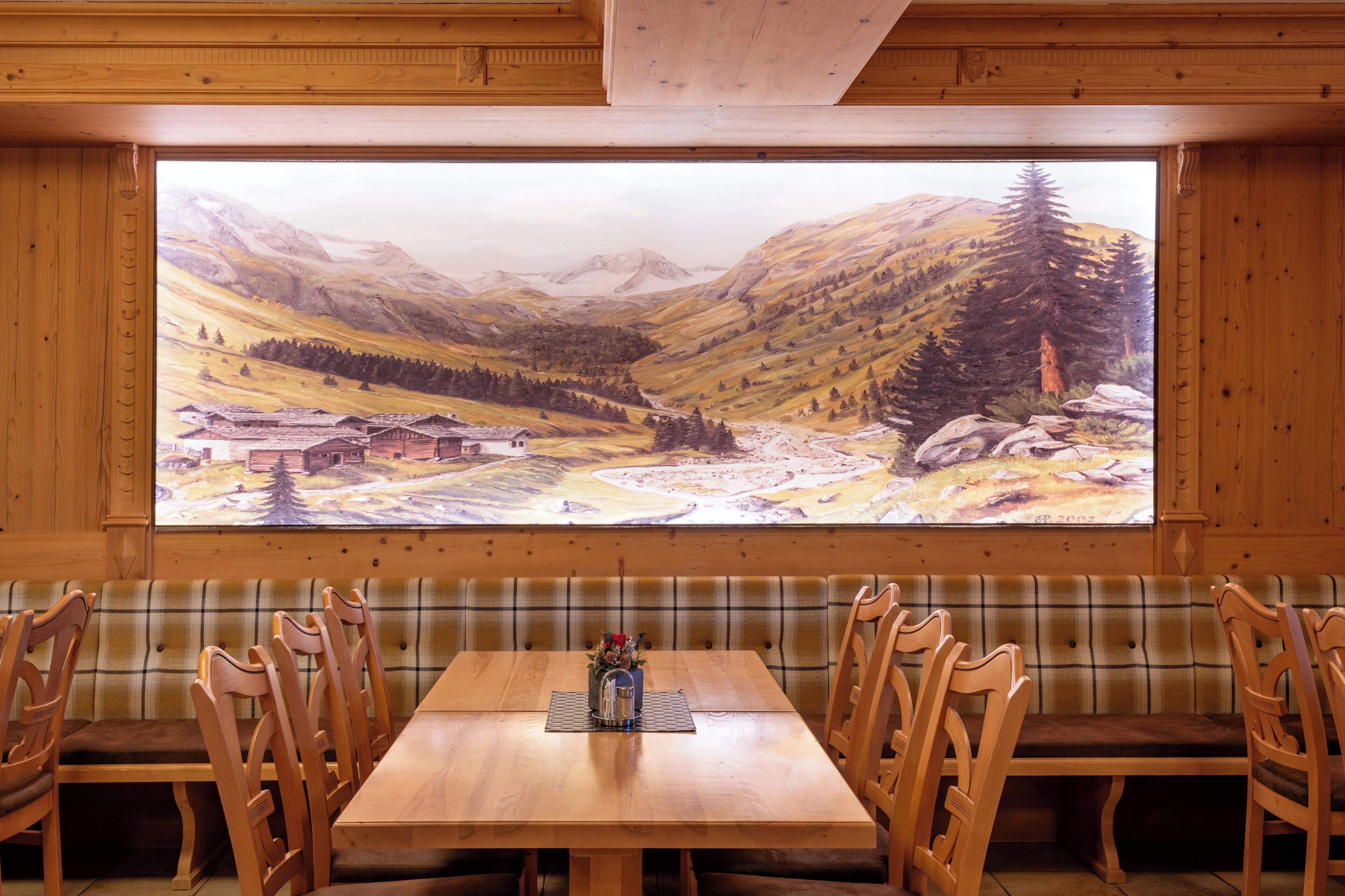 Ein Essbereich mit Holzböden, an der Wand ein großes Gemälde einer Berglandschaft, einem Holztisch und karierten Bänken.