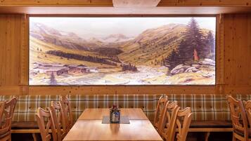 Ein Essbereich mit Holzböden, an der Wand ein großes Gemälde einer Berglandschaft, einem Holztisch und karierten Bänken.