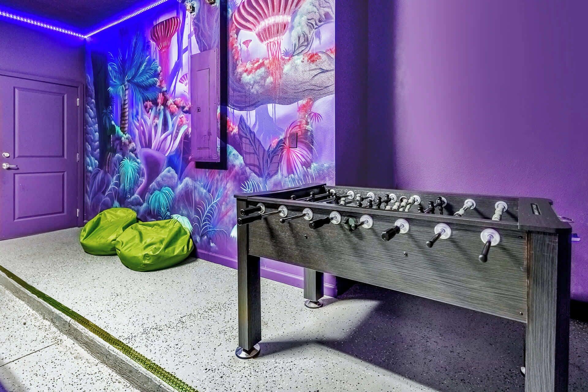A room with a foosball table, green bean bags, and a purple wall with a mural.