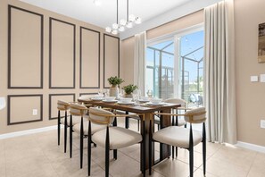 Ein Esszimmer mit einem großen Tisch, gedeckt für sechs Personen, umgeben von sechs Stühlen mit gepolsterten Sitzen und schwarzen Metallbeinen. Der Raum ist modern gestaltet, mit beigen Wänden, einem großen Fenster mit Vorhängen und einer Kristallleuchter.