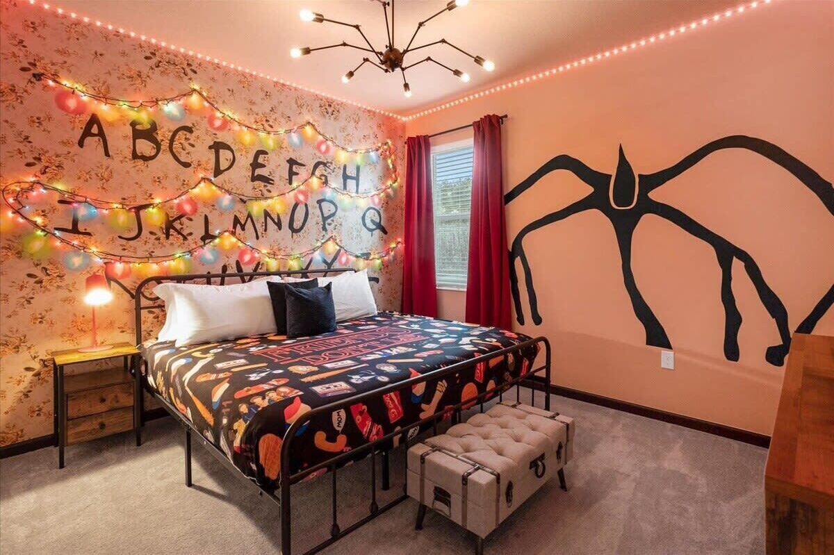 A bedroom with a bed, a bedside table, a lamp, a wall with a decorative string of lights spelling out the alphabet, and a large black abstract wall art piece.