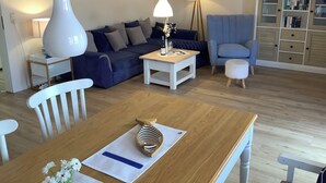 A living room with a sofa, a coffee table, a lamp, a chair, a small table with a bottle and glasses, and a cabinet with books.