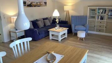 Ein Wohnzimmer mit einer Couch, einem Couchtisch, einer Lampe, einem Sessel, einem kleinen Tisch mit einer Flasche und Gläsern sowie einem Schrank mit Büchern.