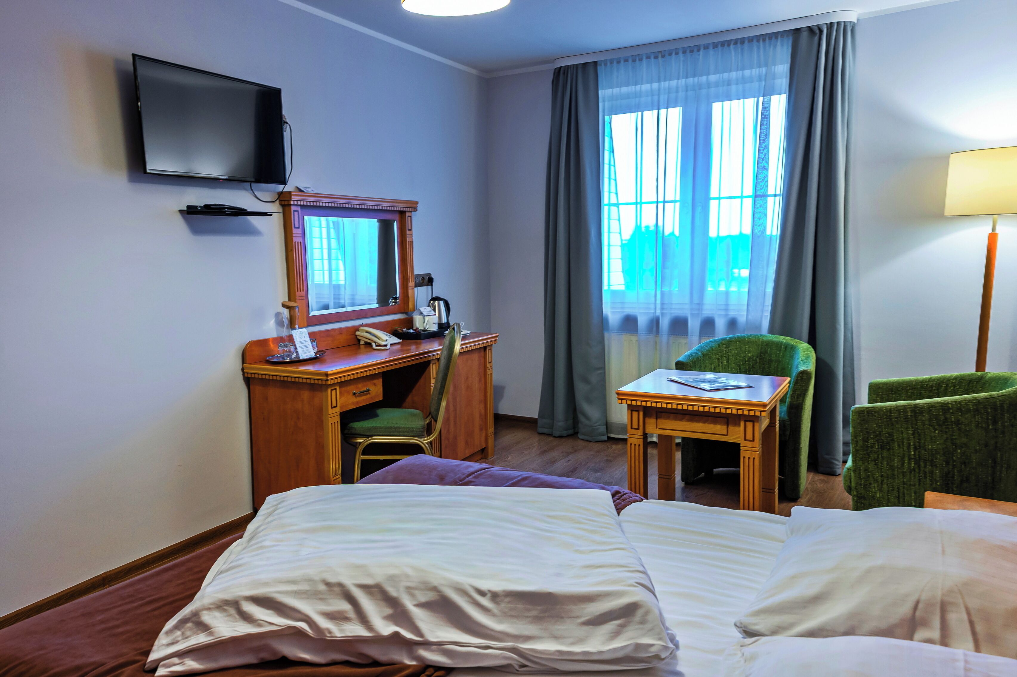 A hotel room with two beds, a desk with a telephone, a chair, a television mounted on the wall, and a window with curtains.