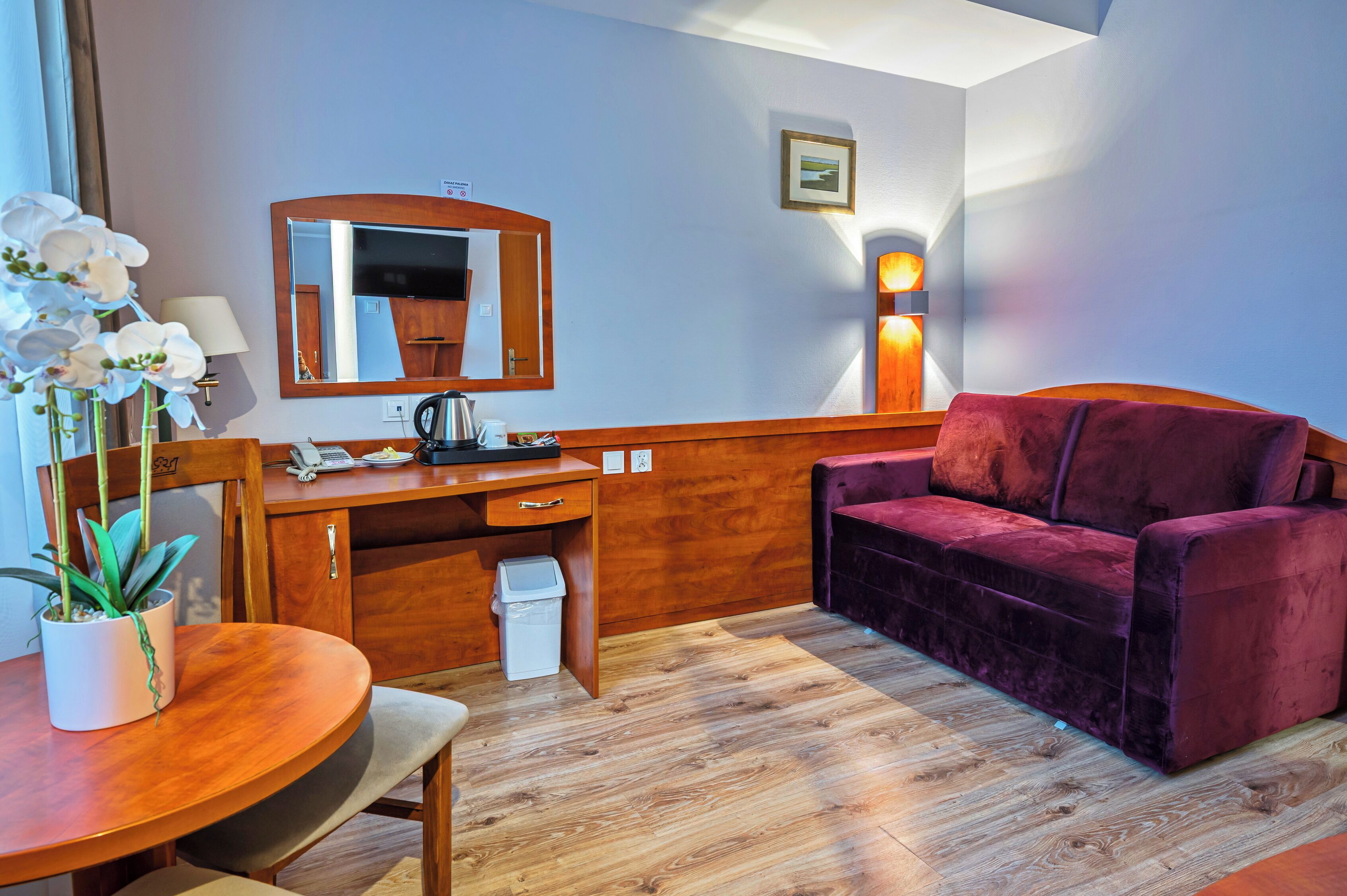 A compact room with a wooden desk, a purple sofa, a small table with a potted plant, a mirror, and a wall-mounted lamp.