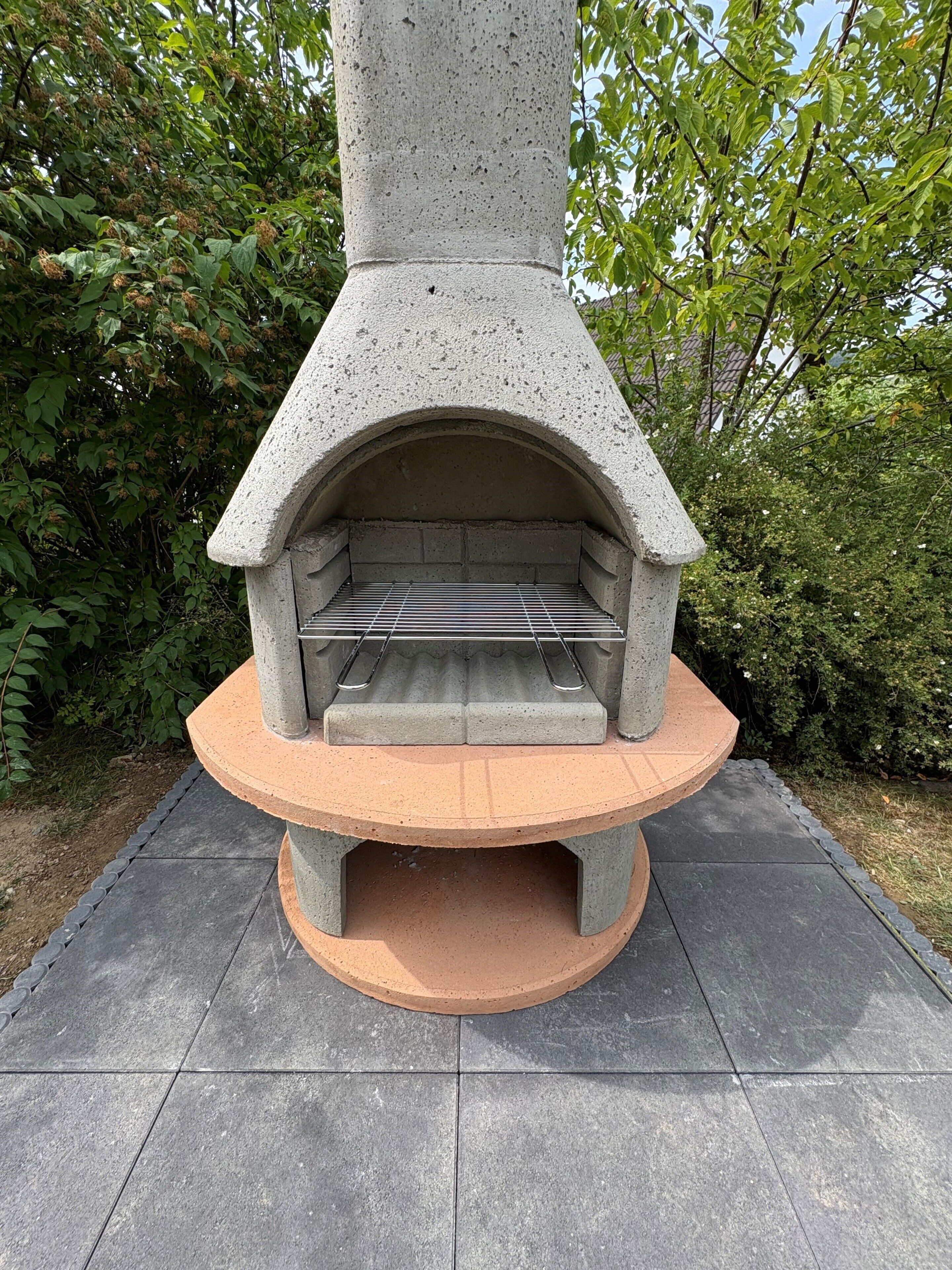 Un four à pizza extérieur en pierre, doté d’une grille métallique à l’intérieur, entouré d’une zone pavée et de verdure.