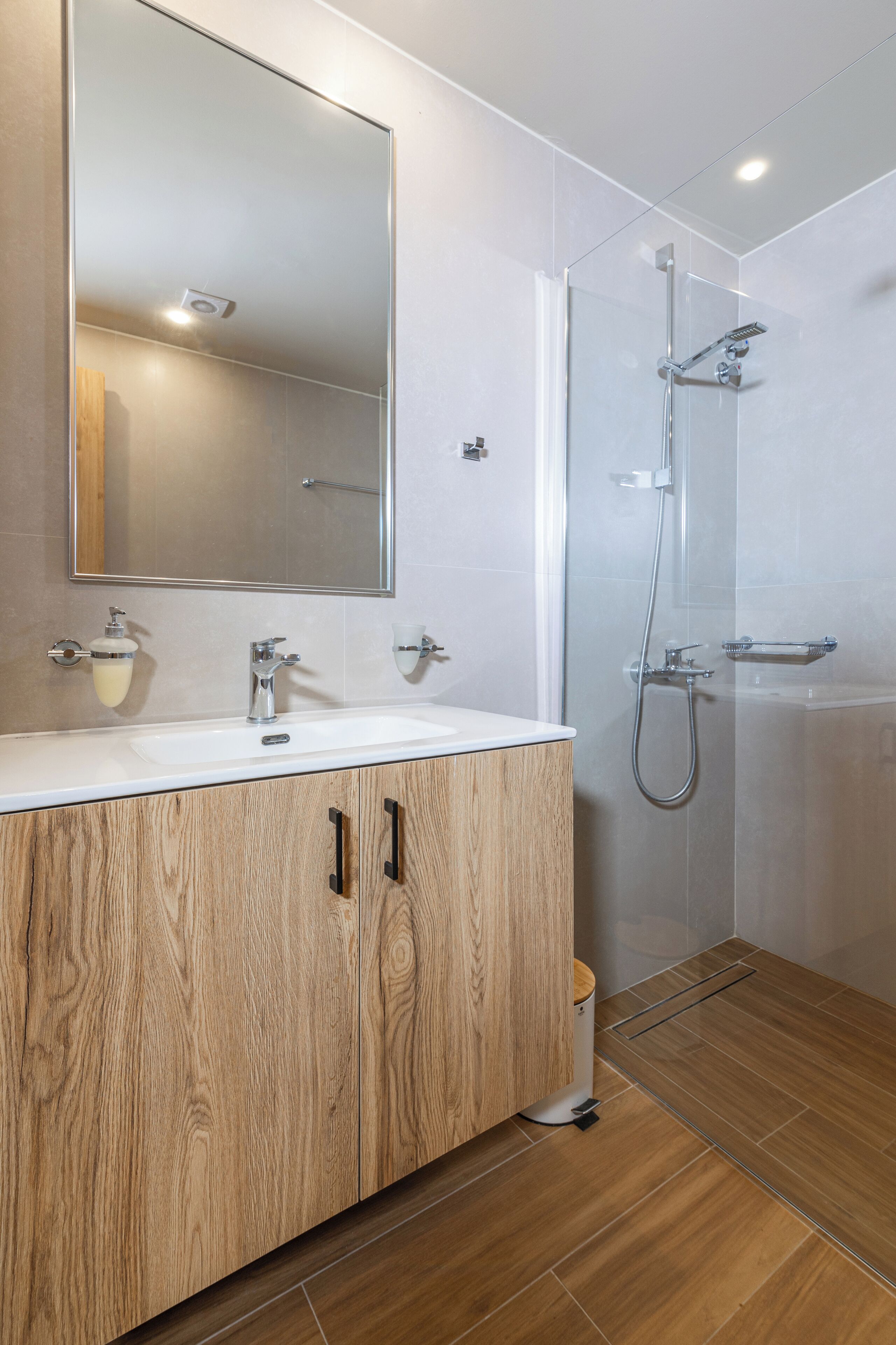 A modern bathroom with a wooden vanity, a large mirror, a shower enclosure, and a white sink.