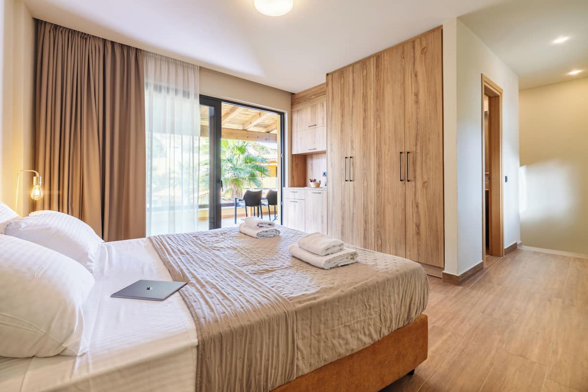 A modern hotel room with a large bed, a laptop on the bed, and a view of the outdoors through the window.