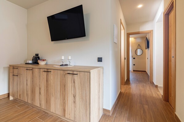 A modern kitchen with a wooden cabinet, a flat-screen TV mounted above the counter, and a hallway leading to a bedroom. - Doce Dioses (Malevizi)