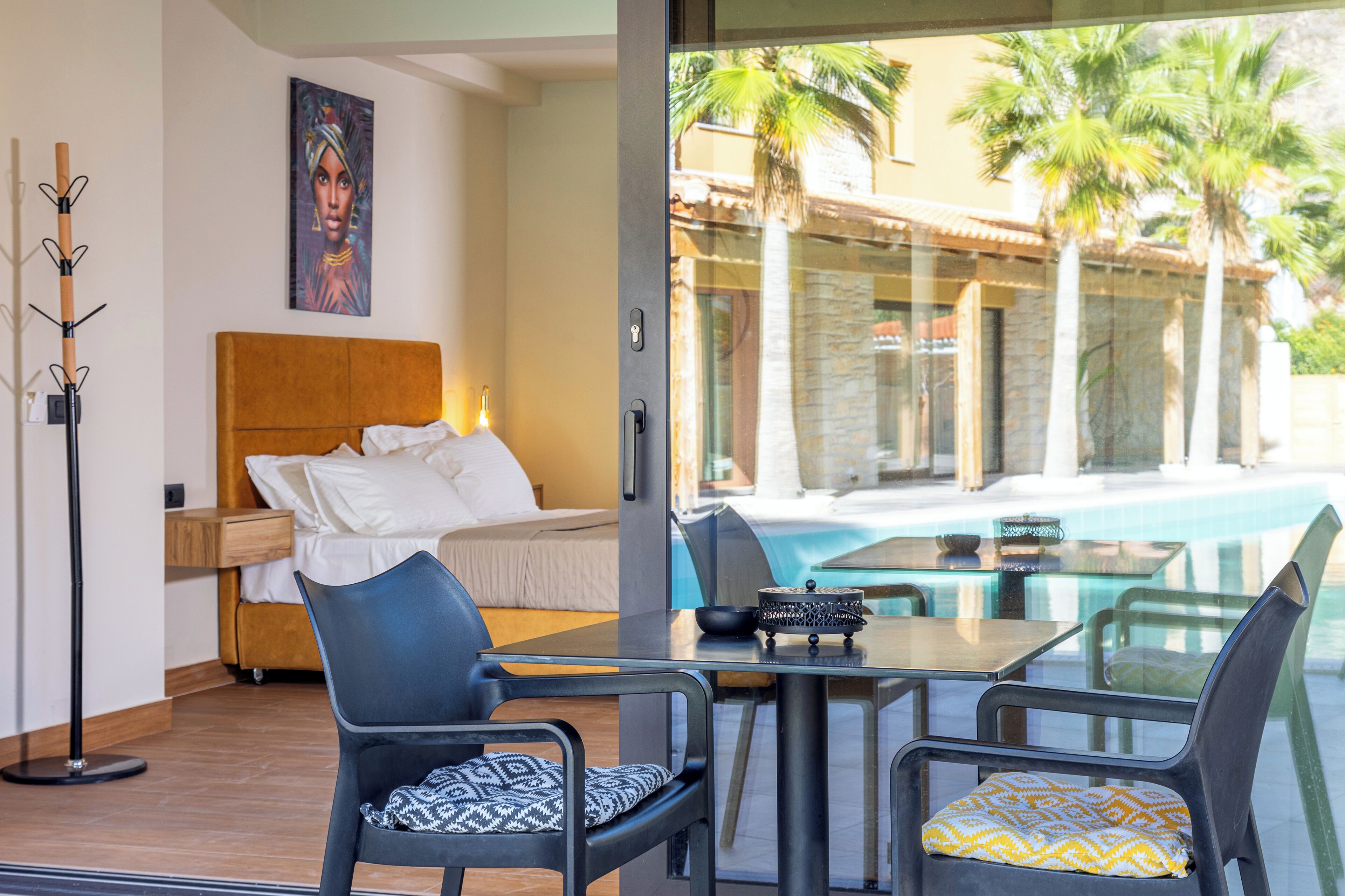 A hotel room with a bed, a glass table, two chairs, a floor lamp, and a view of a swimming pool with palm trees.