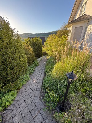 Un chemin pavé mène à un bâtiment, lequel est entouré d’une végétation luxuriante et se dresse sous un ciel dégagé.