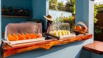Montaje de bufé con una tabla de madera, una cubierta de plástico transparente sobre un plato de fruta cortada y una cesta de naranjas.