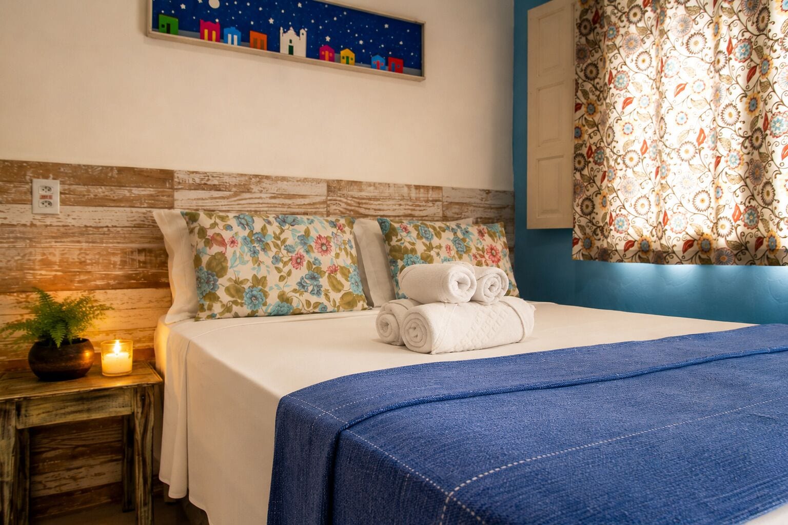 A neatly made bed with a blue blanket, floral pillows, and rolled towels on a wooden nightstand with a lit candle.