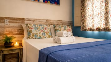 A neatly made bed with a blue blanket, floral pillows, and rolled towels on a wooden nightstand with a lit candle.