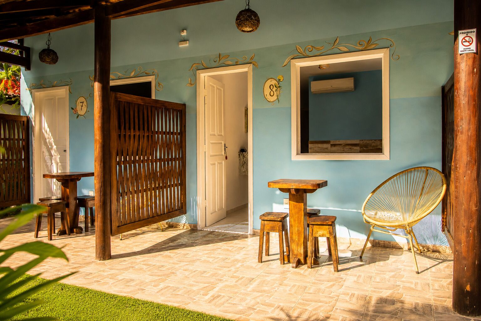 A covered outdoor seating area with a wooden table and chairs, a wicker chair, and a door with a number sign.