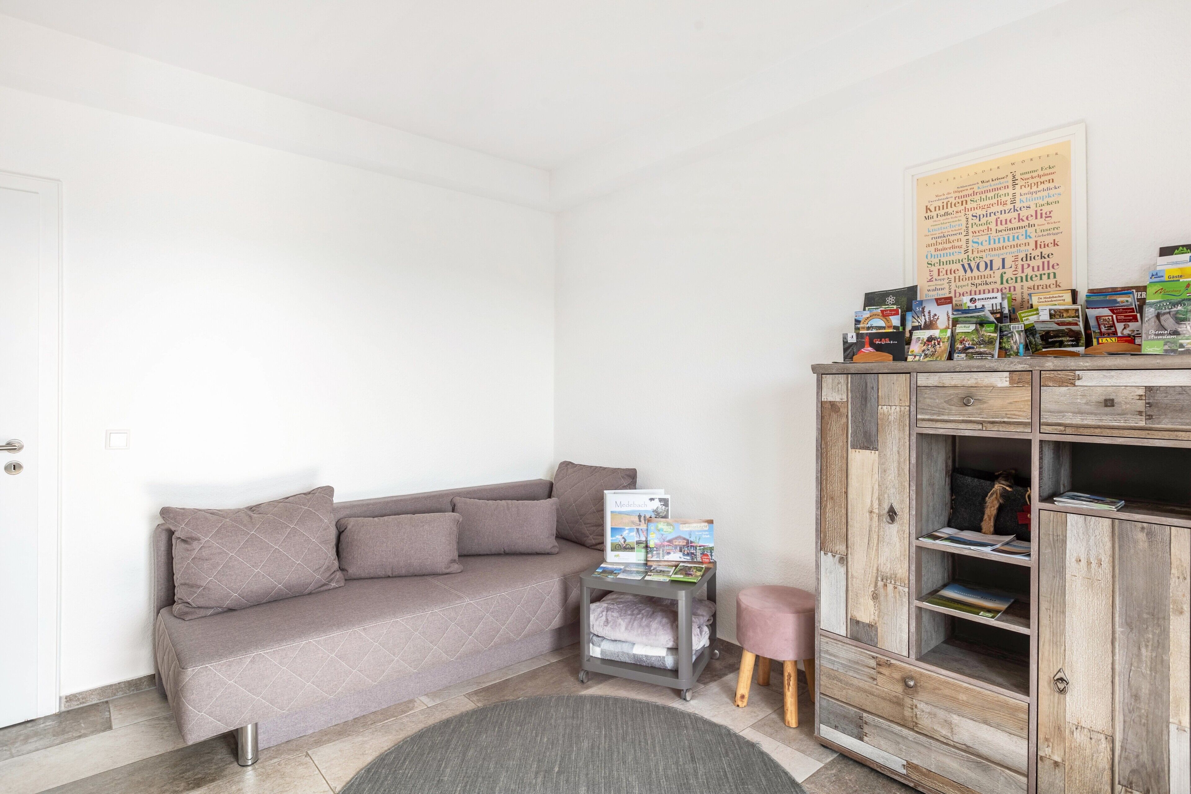 A living room with a grey sofa, a small table with magazines, a wooden cabinet, and a framed word cloud on the wall.