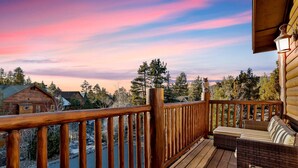 Eine Holzterrasse mit Geländer, ein Korbsessel und Blick auf ein bewaldetes Gebiet mit Häusern im Hintergrund.