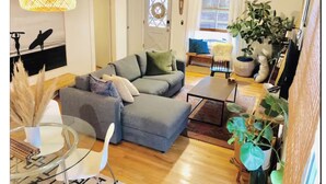 A living room with a grey sectional sofa, a glass coffee table, and a white chair. There is a plant on the right and a painting on the wall. - Hip & Historic downtown Vancouver bungalow (Vancouver)