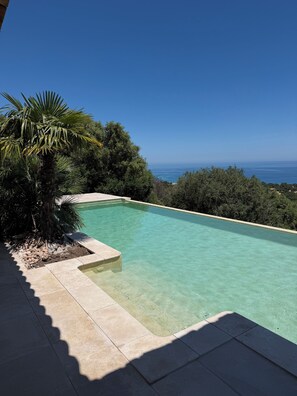 Ein klares Schwimmbecken mit Blick auf das Meer, umgeben von viel Grün und einer Steindeck.