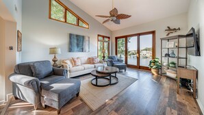 Un salon spacieux avec un canapé gris, deux fauteuils, une table basse et un ventilateur de plafond. De grandes fenêtres laissent entrer la lumière naturelle et offrent une vue sur l’extérieur.