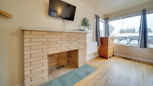 Een woonkamer met een metselwerk open haard, een tv aan de muur, een raam met gordijnen en uitzicht op buiten.