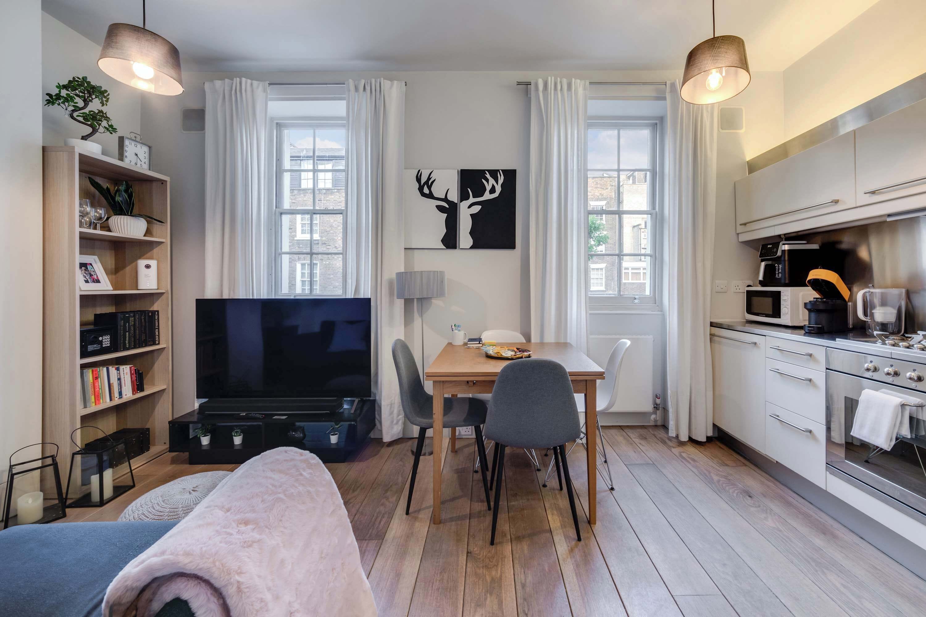 A modern living room with a TV, sofa, and dining area. There is a kitchen with appliances and a dining table.