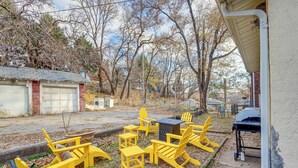 A patio area with yellow Adirondack chairs and a grill, surrounded by trees and a building with a garage. - 3 Mi to Dtwn: Omaha Escape w/ Game Room & Patio! (Omaha)