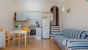 Een compacte keuken met witte kastjes, een eettafel met stoelen, een magnetron, een koelkast en een gestreept bankje.