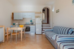 A compact kitchen with white cabinets, a dining table with chairs, a microwave, a refrigerator, and a striped sofa. - GuestHost - Cozy 50 square meters villa, perfect for four people, with direct access from the garden.The property is just a 12-minute drive from Porto Giunco ​​beach and a 15-minute drive from Capo Carbonara beach.The accommodation comprises a living (Villasimius)