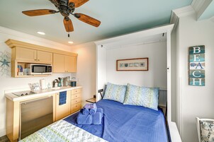 Un dormitorio pequeño con cama, ventilador de techo, kitchenette y una obra de arte colgada en la pared.
