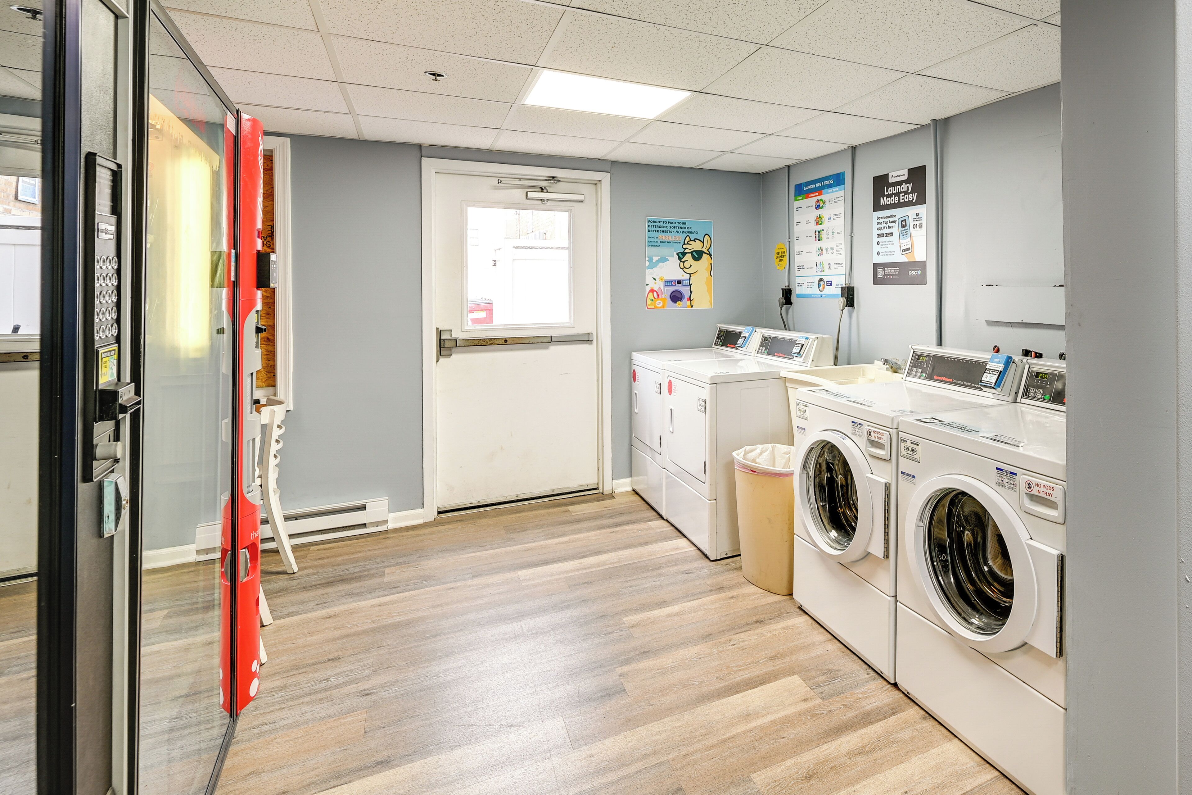 A laundry room with two front-loading washers and dryers, a trash bin, and a door leading to another area.