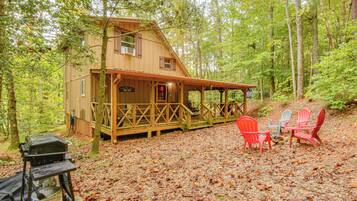 Une cabane avec une véranda, des fauteuils Adirondack rouges et gris, et un barbecue.