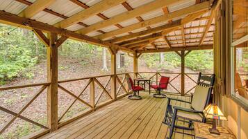 Una terraza de madera con techo metálico, rodeada de árboles. Hay dos mecedoras y una mesa con una lámpara.