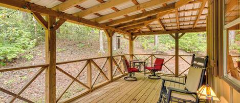 A wooden deck with a metal roof, surrounded by trees. There are two rocking chairs and a table with a lamp.